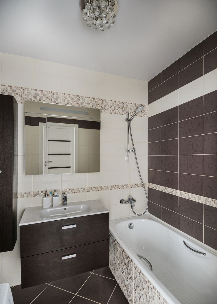Törtfehér és szürkésbarna padló és falburkolatok, utóbbi mintás és mozaik elemekkel, barna bútor fürdőkád
