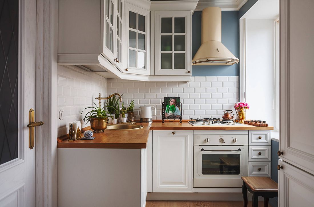 Kis konyha fehér konyhabútorral, felül üvegezett szekrényekkel, meleg fa munkalappal és padlóval, kék faldekorációval, fehér csempe hátfallal