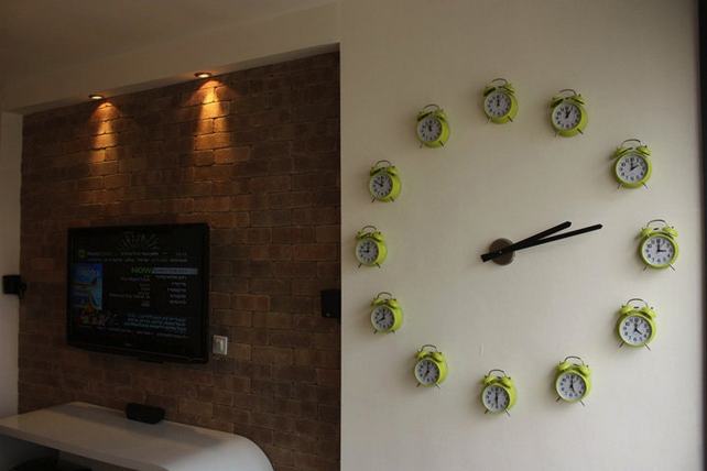 egy ötletes és hatalmas óra a falon, melynek számait az adott órára beállított öreg vekkerek adják