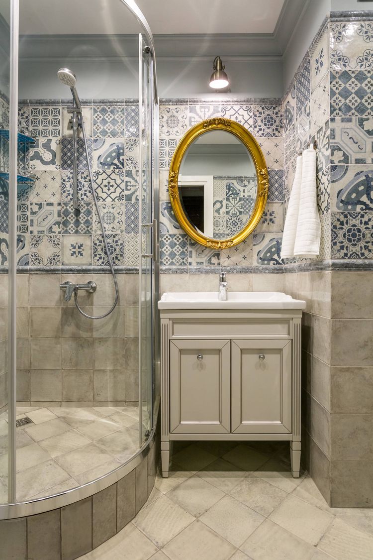 Szürkés-bézs burkolat padlón és a falak alsó részén, mintás patchwork burkolat felül, szürke festett sáv a mennyezet alatt, zuhanyfülke, aranyszínű tükörkeret